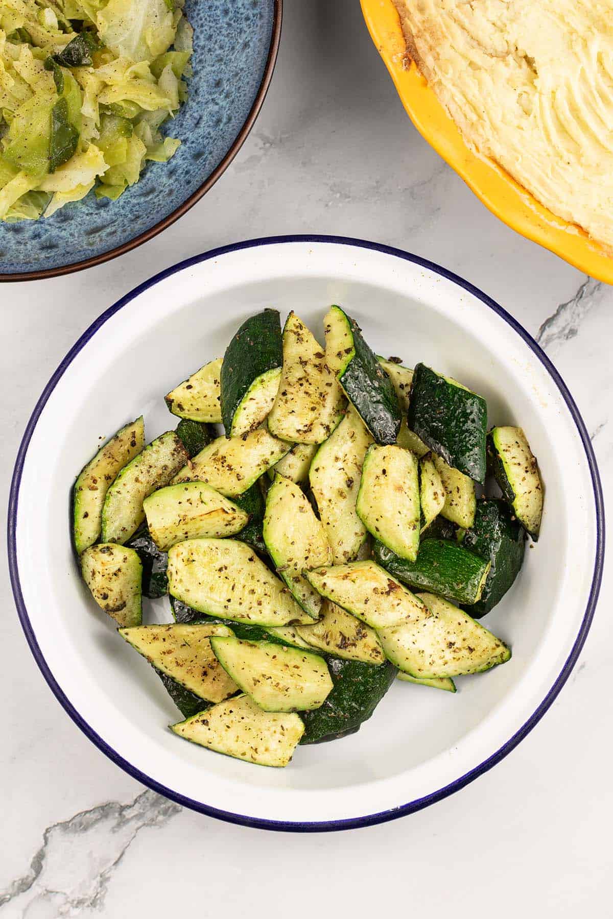 Easy air fryer courgettes in a round blue rimmed white enamel dish with a bowl of cabbage and a pie in the background.