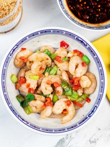 Easy salt and chilli pepper prawns in a blue and white oriental bowl, with a bowl of dipping sauce and jar of Chinese seasoning in the background.