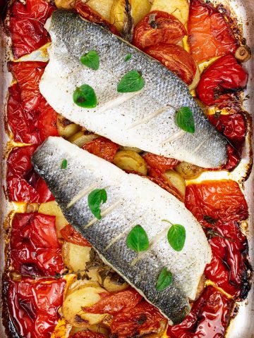 Close up of two portions of easy sea bass with roasted peppers and potatoes in a roasting tin.