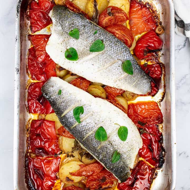 Close up of two portions of easy sea bass with roasted peppers and potatoes in a roasting tin.