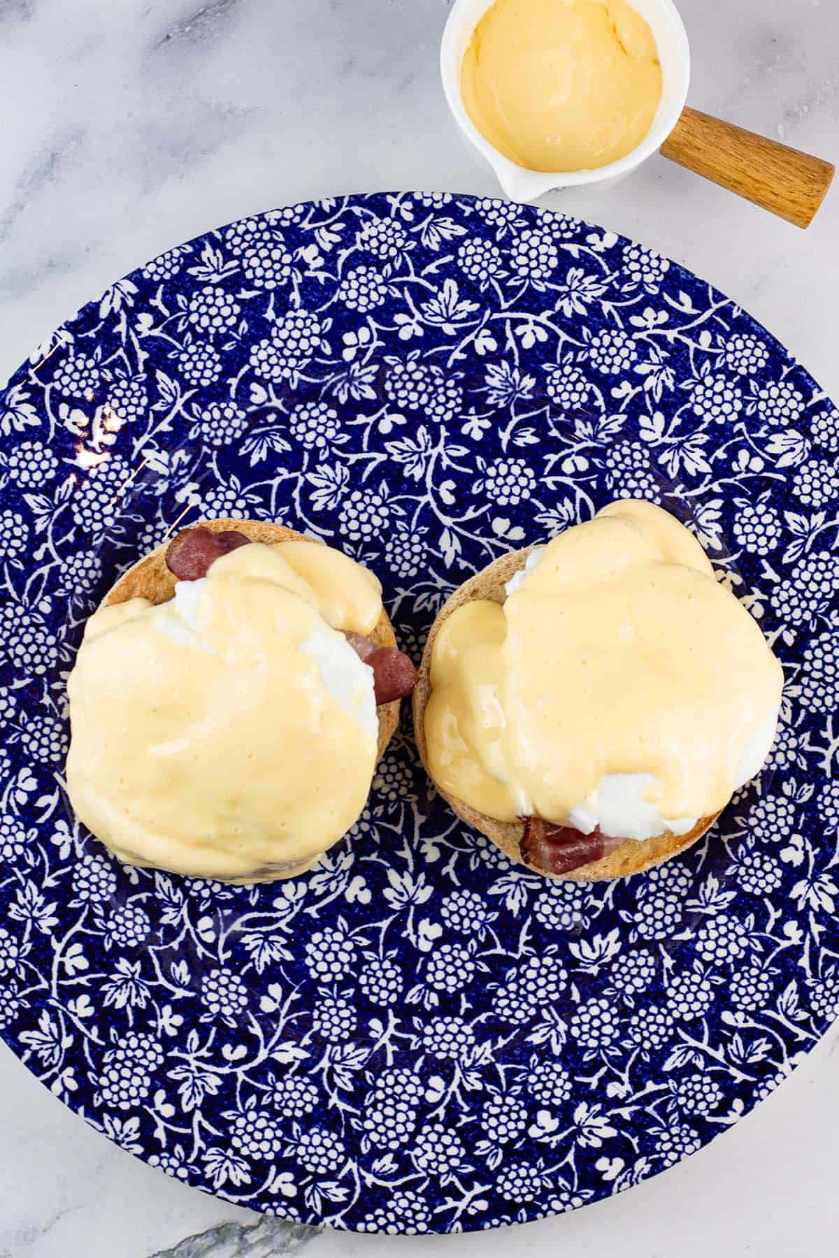 Eggs benedict with quick hollandaise sauce on a blue and white patterned dinner plate, with a small jug of sauce on the side.