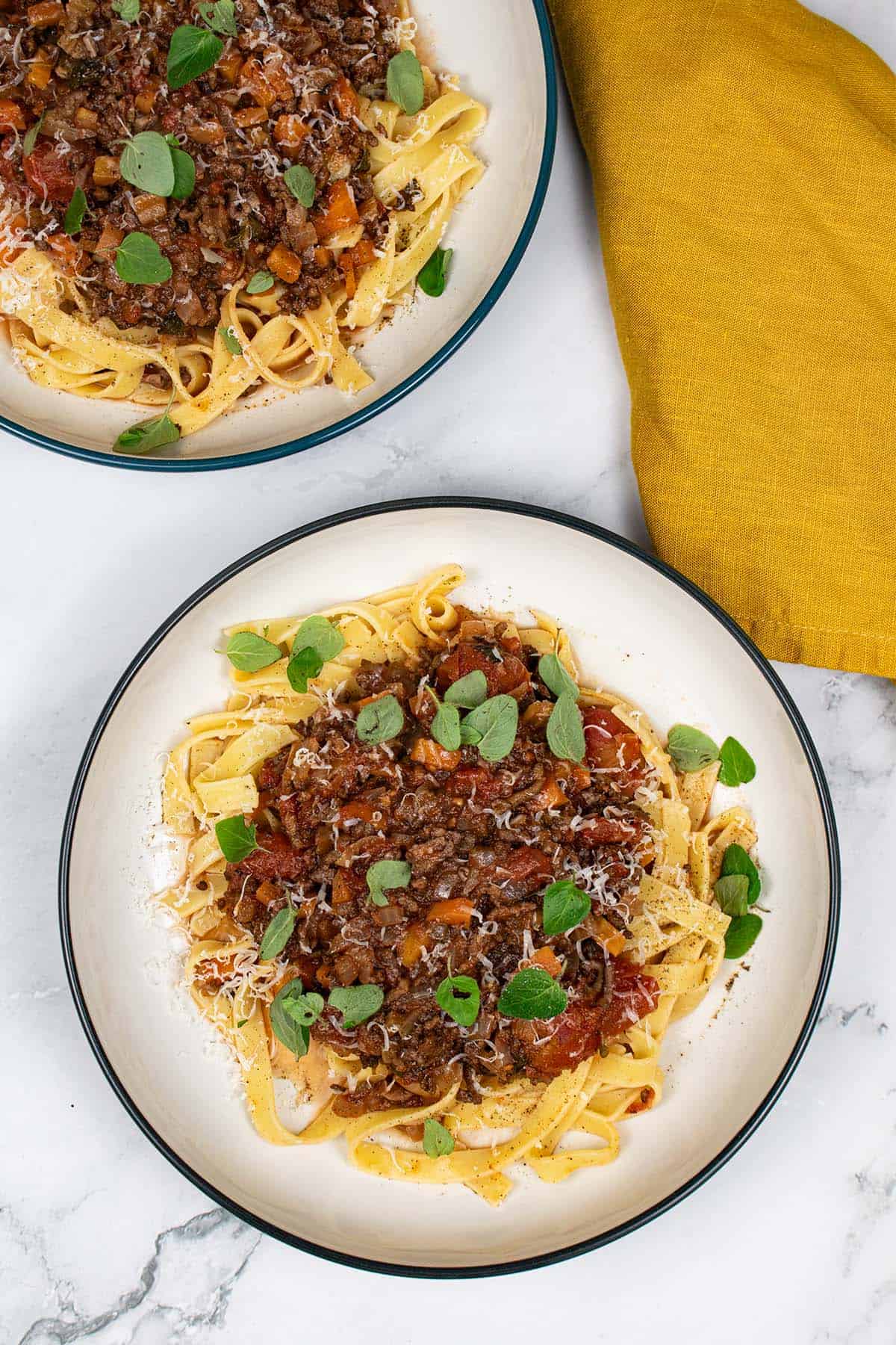 2 pasta bowls of lamb, tomato and oregano ragu with tagliatelle and a mustard napkin on the side.