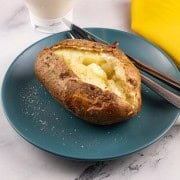 Perfect air fryer baked potato on a teal side plate seasoned with salt and pepper and a knob of butter. A fork, knife, yellow napkin and bottle of mayo on the side.