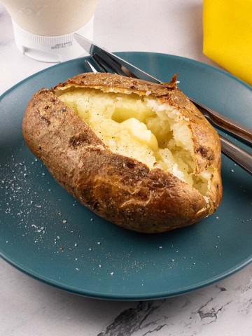 Perfect air fryer baked potato on a teal side plate seasoned with salt and pepper and a knob of butter. A fork, knife, yellow napkin and bottle of mayo on the side.