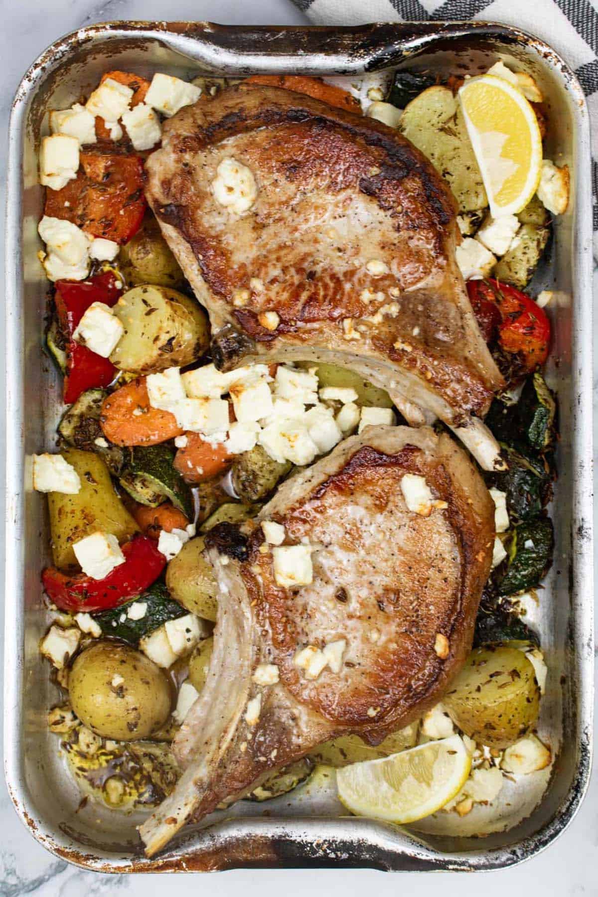 Pork chops with roast vegetables and feta in a roasting tin with a black and white checked towel on the side.
