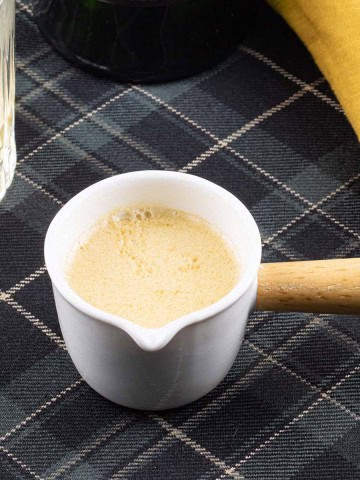 Whisky cream sauce in a cream jug with wooden handle sitting on a tartan cloth with a glass of whisky and bottle in the background.