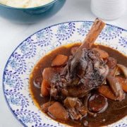 Rioja-braised lamb shank with chorizo and garlic on a blue and white patterned dinner plate, with a bowl of mashed potatoes and small jug of sauce on the side.