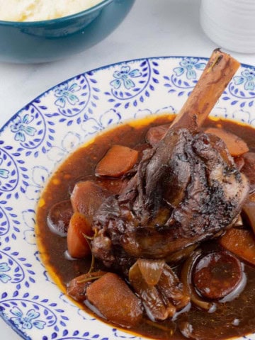 Rioja-braised lamb shank with chorizo and garlic on a blue and white patterned dinner plate, with a bowl of mashed potatoes and small jug of sauce on the side.