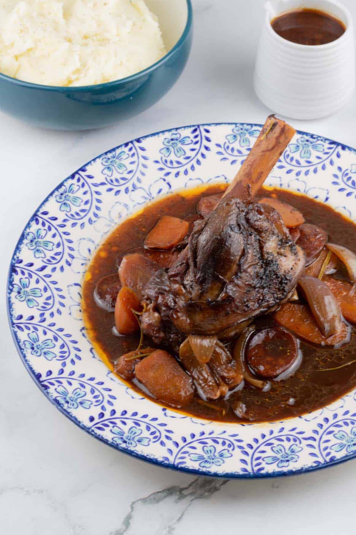 Rioja-braised lamb shank with chorizo and garlic on a blue and white patterned dinner plate, with a bowl of mashed potatoes and small jug of sauce on the side.