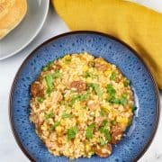 A portion of sausage and pea risotto made in the Thermomix in a blue patterned bowl with a crusty bread roll and mustard napkin on the side.