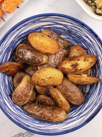 Sautéed new potatoes with rosemary in a blue and white striped bowl, with dishes of za'atar carrots and tenderstem broccoli on the side.