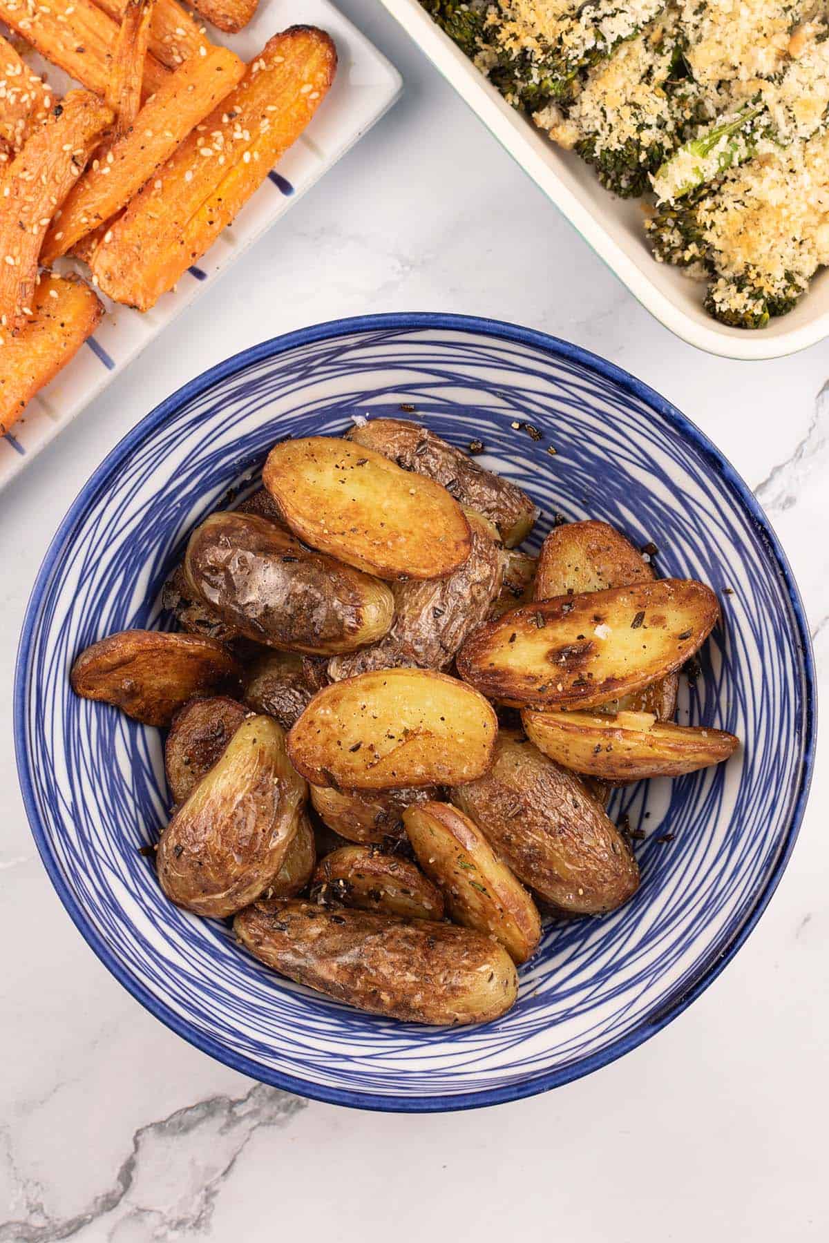 Sautéed new potatoes with rosemary in a blue and white striped bowl, with dishes of za'atar carrots and tenderstem broccoli on the side.