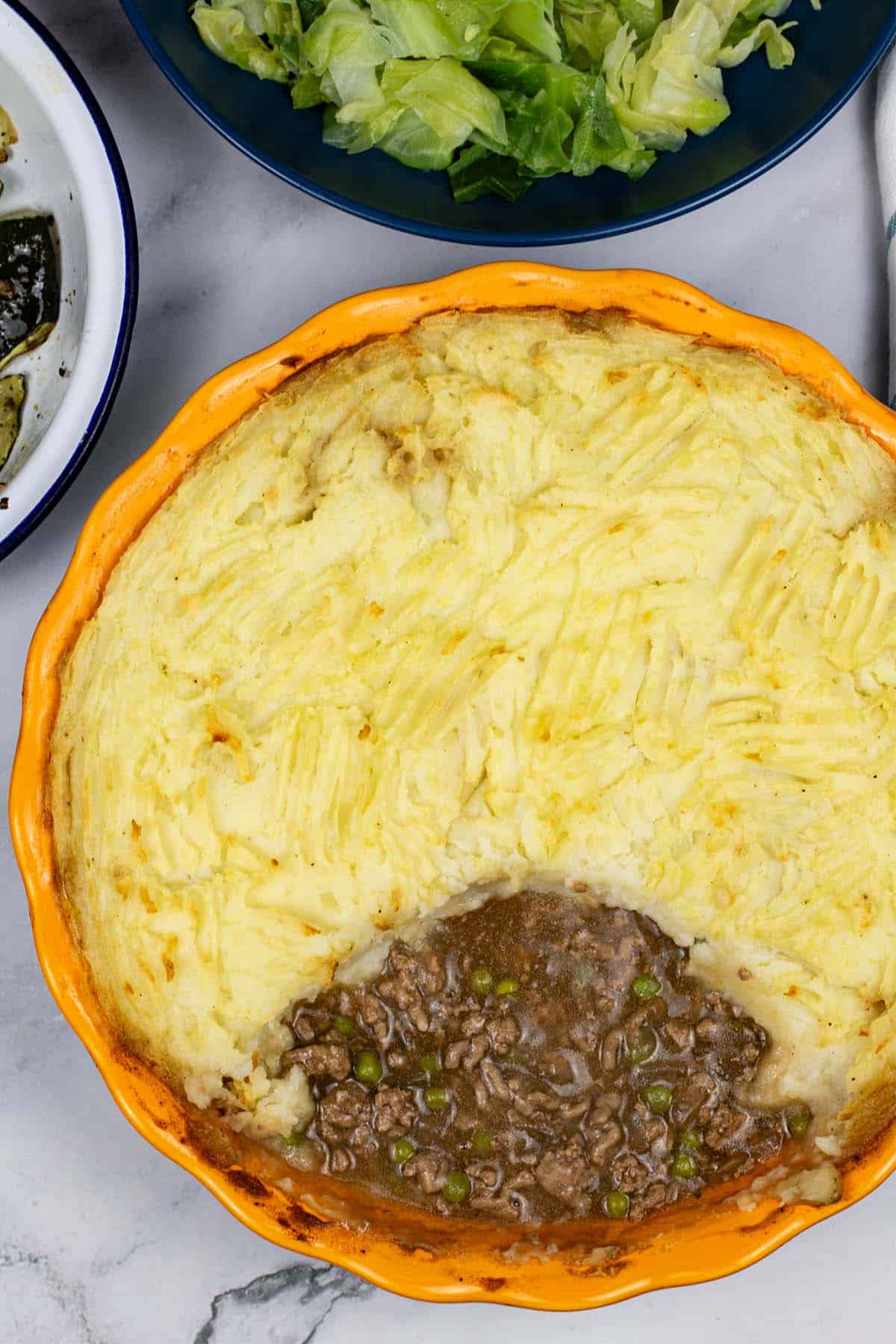 Shepherd's pie in a round yellow and blue pie dish with a portion of potatoes removed, and a bowl of buttered cabbage in the background.