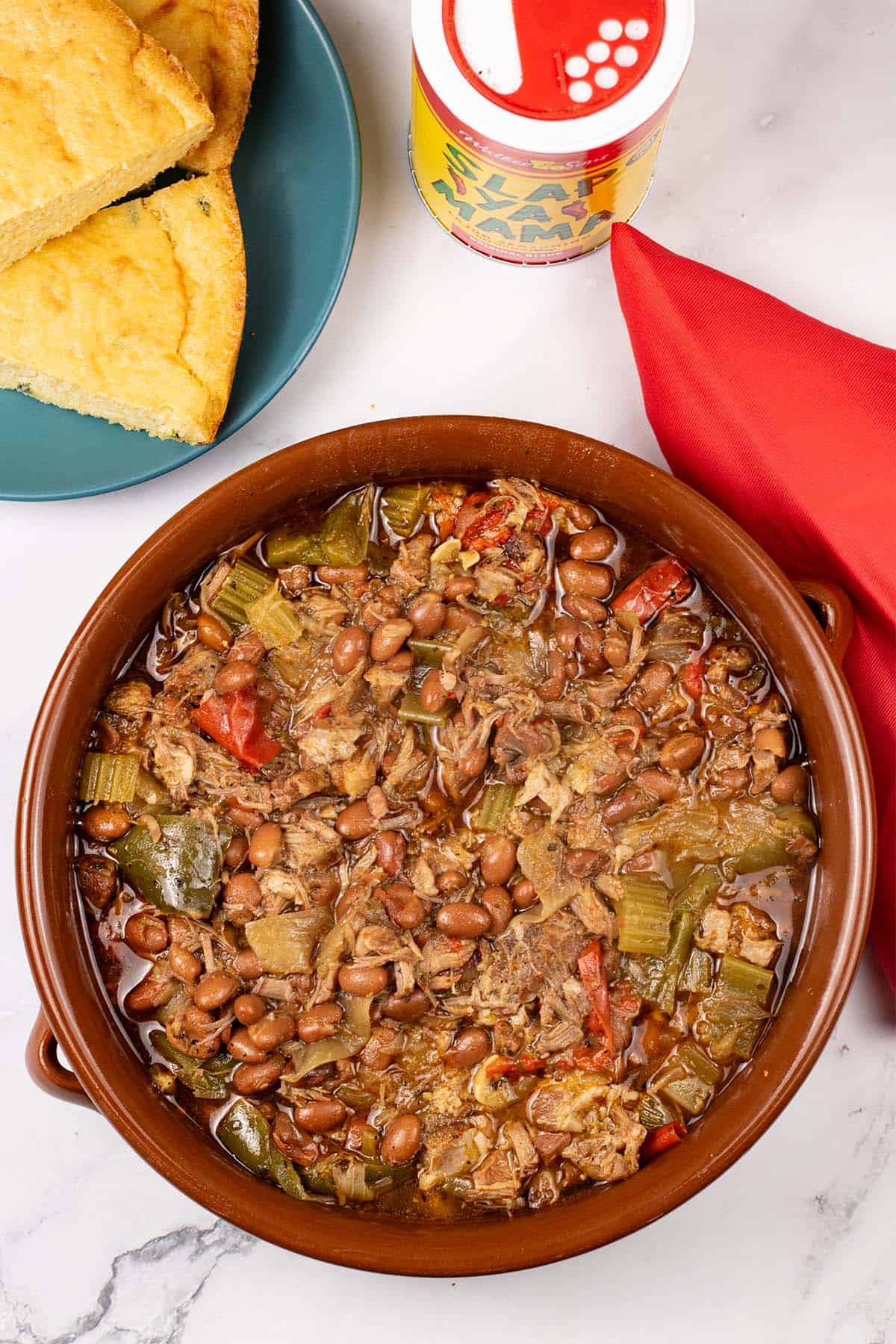 Slow cooker cajun pork and red beans in a large round terracotta dish with a plate of cornbread, tub of cajun seasoning and red napkin on the side.