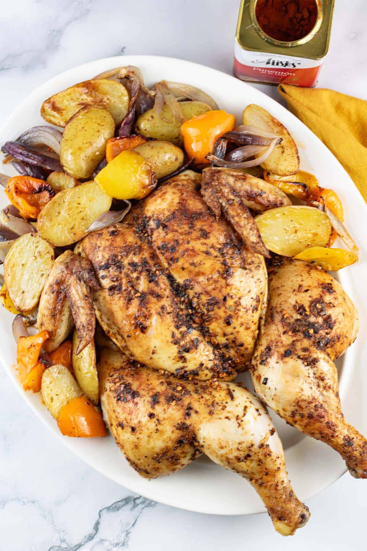 Spanish spatchcock chicken with vegetables on a white platter with tin of hot smoked pimenton and a mustard napkin on the side.