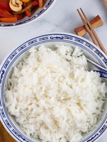 Steamed jasmine rice in a blue and white oriental bowl with spoon, chopsticks and a bowl of chicken and cashew nut stir-fry on the side.
