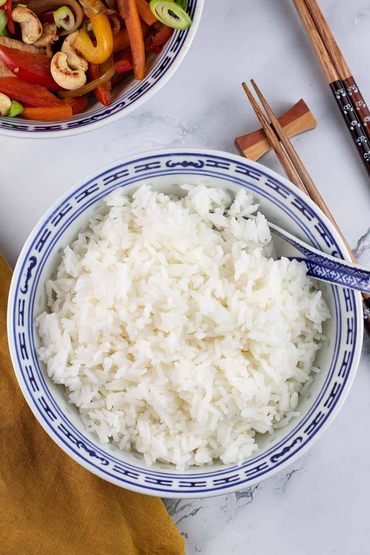 Steamed jasmine rice in a blue and white oriental bowl with spoon, chopsticks and a bowl of chicken and cashew nut stir-fry on the side.