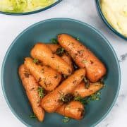 Thyme butter roasted Chantenay carrots in a teal bowl, with bowls of cabbage and mashed potatoes in the background.