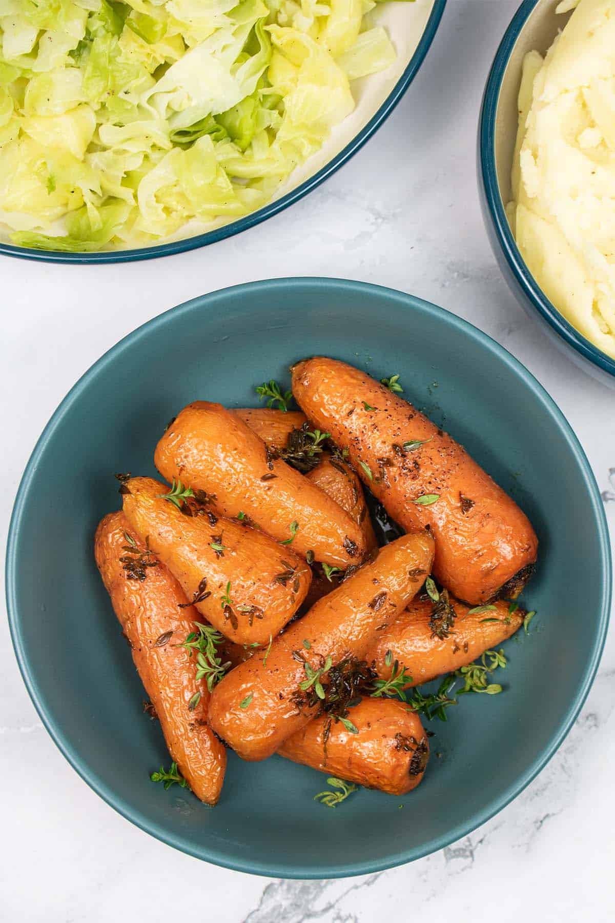 Thyme butter roasted Chantenay carrots in a teal bowl, with bowls of cabbage and mashed potatoes in the background.