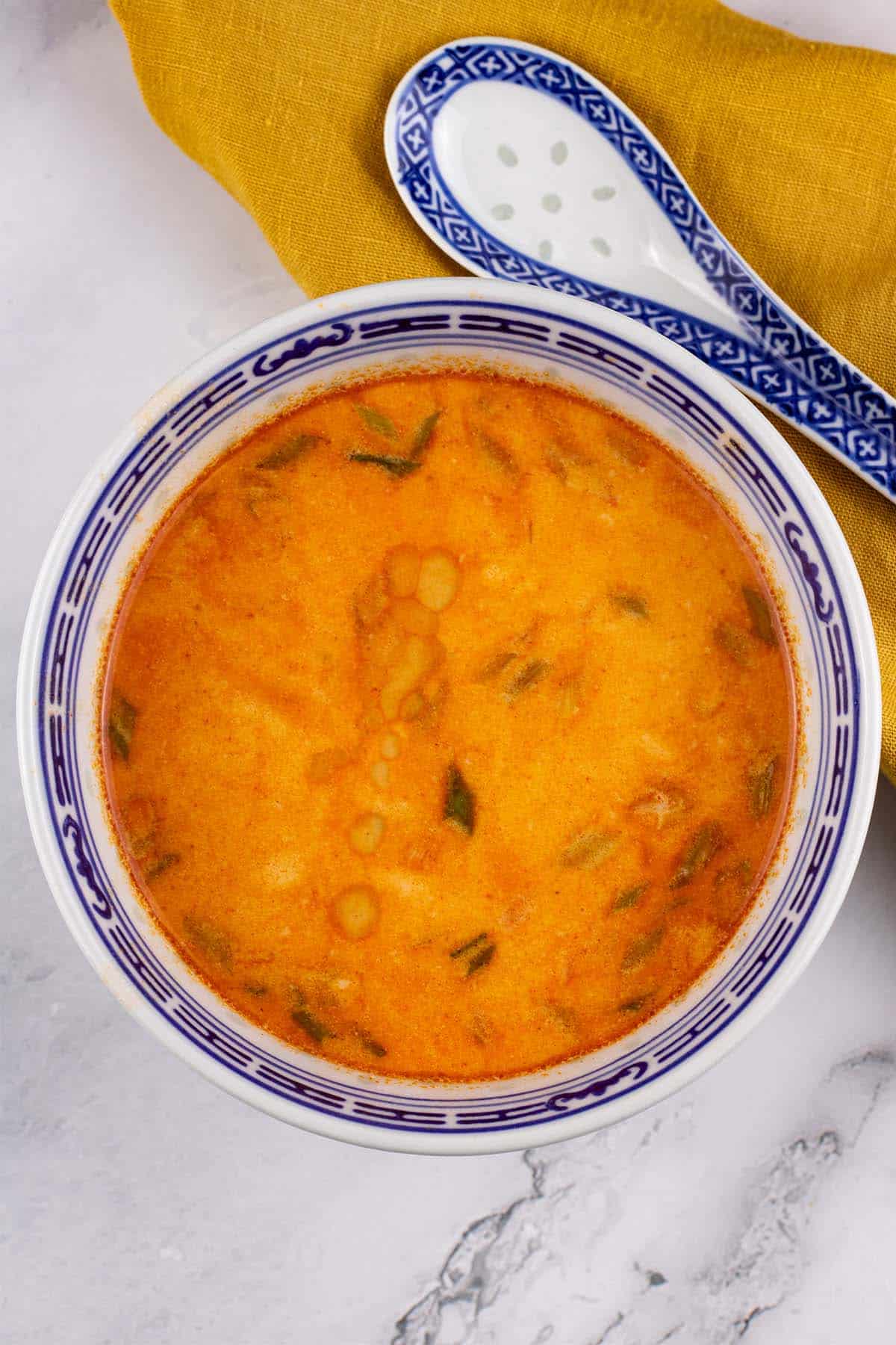 Tom yum soup in a small black blue and white oriental bowl with a spoon and mustard napkin on the side.