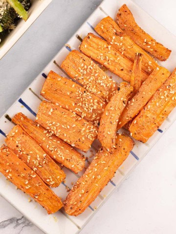 Za'atar roasted carrots on a rectangle striped platter with a dish of tenderstem broccoli on the side.