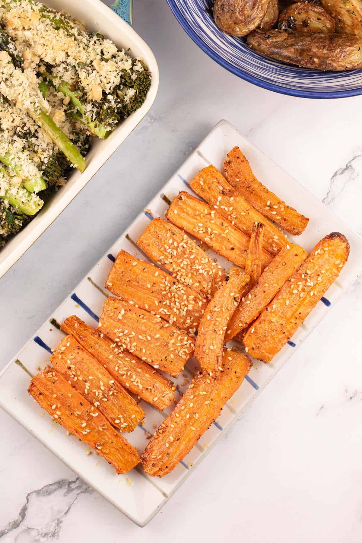 Za'atar roasted carrots on a rectangle striped platter with a dish of tenderstem broccoli and bowl of sautéed potatoes on the side.