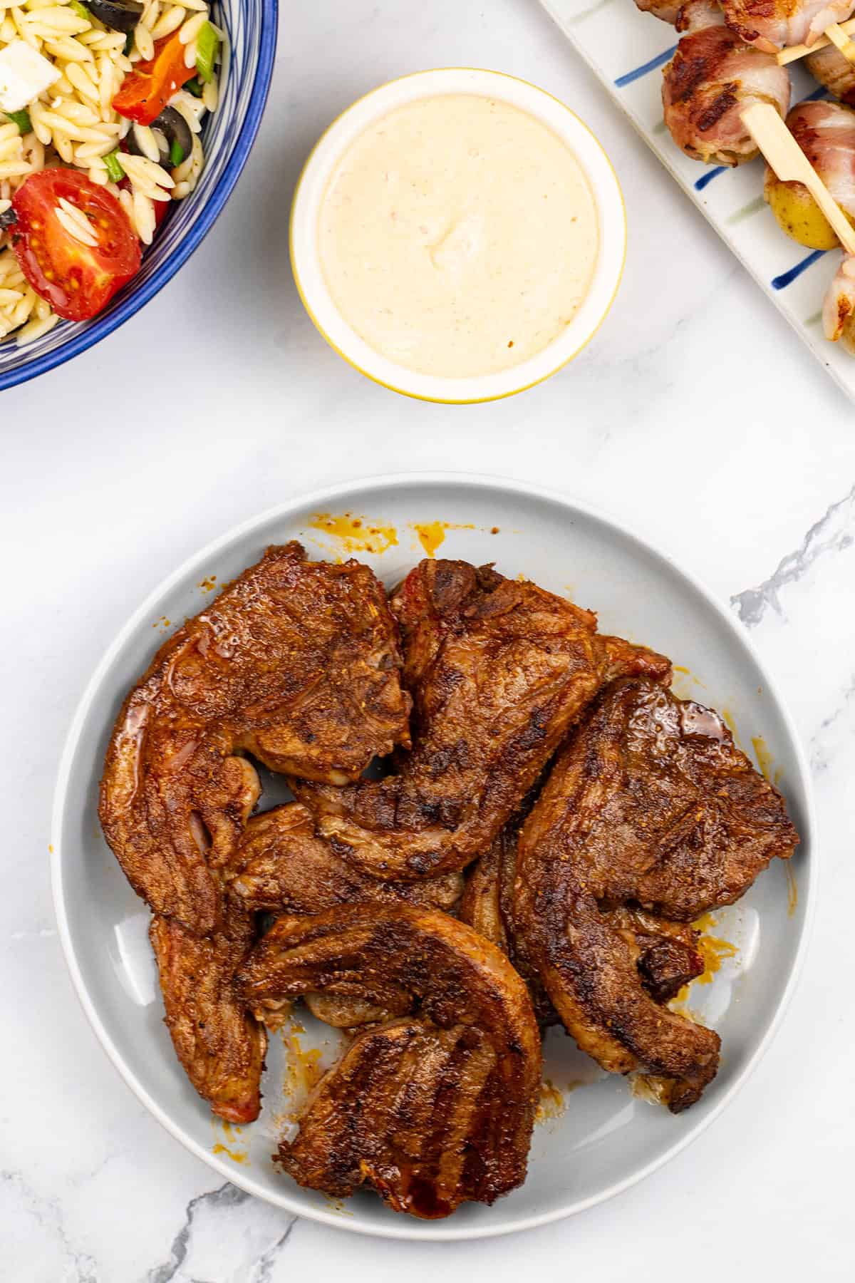 BBQ merguez lamb chops on a grey plate with a bowl of orzo salad with feta, and a plate of baby new potatoes wrapped in bacon with harissa dip on the side.