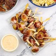 Baby new potatoes wrapped in bacon on a striped platter with harissa cheese dip in a yellow ramekin. A bowl of orzo salad with feta and a plate of BBQ merguez lamb chops on the side.