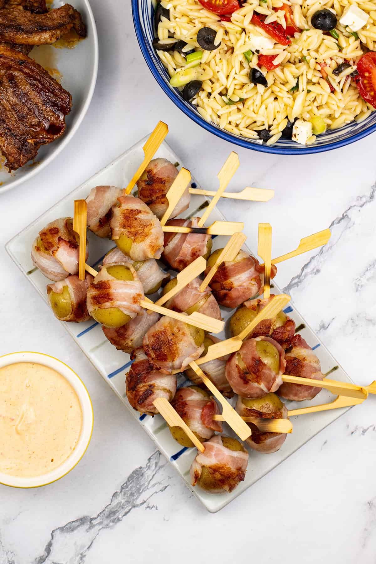 Baby new potatoes wrapped in bacon on a striped platter with harissa cheese dip in a yellow ramekin. A bowl of orzo salad with feta and a plate of BBQ merguez lamb chops on the side.