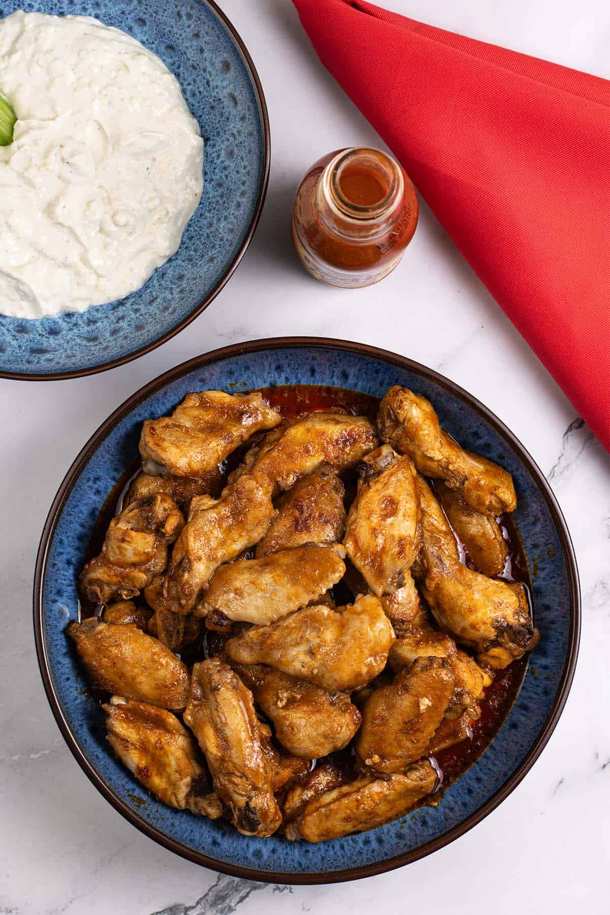 Buffalo chicken wings in a blue patterned bowl with a bowl of blue cheese dip, bottle of Frank's RedHot Wings Buffalo sauce and red napkin on the side.