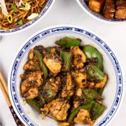 Chicken with green peppers and black bean sauce in an oriental bowl with bowls of fried noodles and Shanghai pork, and chopsticks on the side.