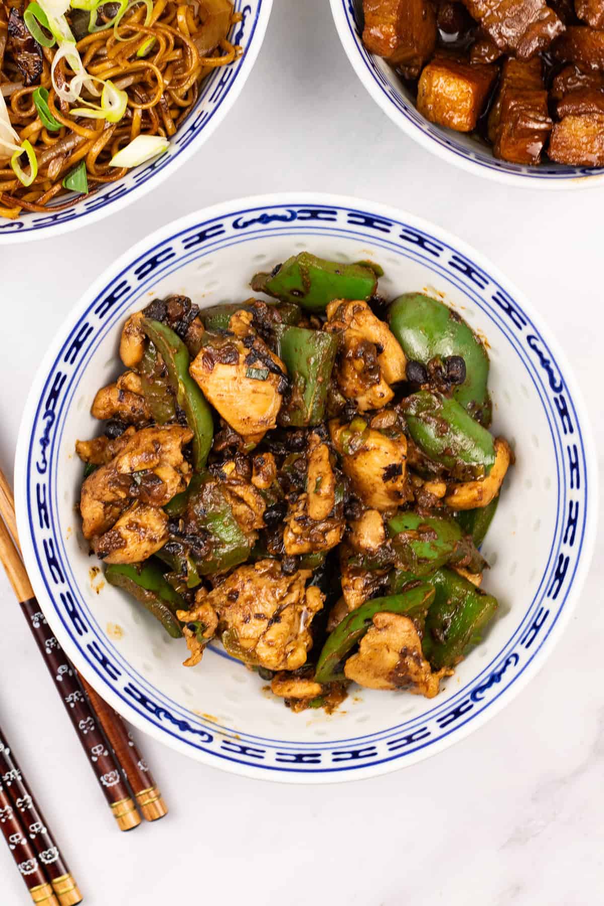 Chicken with green peppers and black bean sauce in an oriental bowl with bowls of fried noodles and Shanghai pork, and chopsticks on the side.