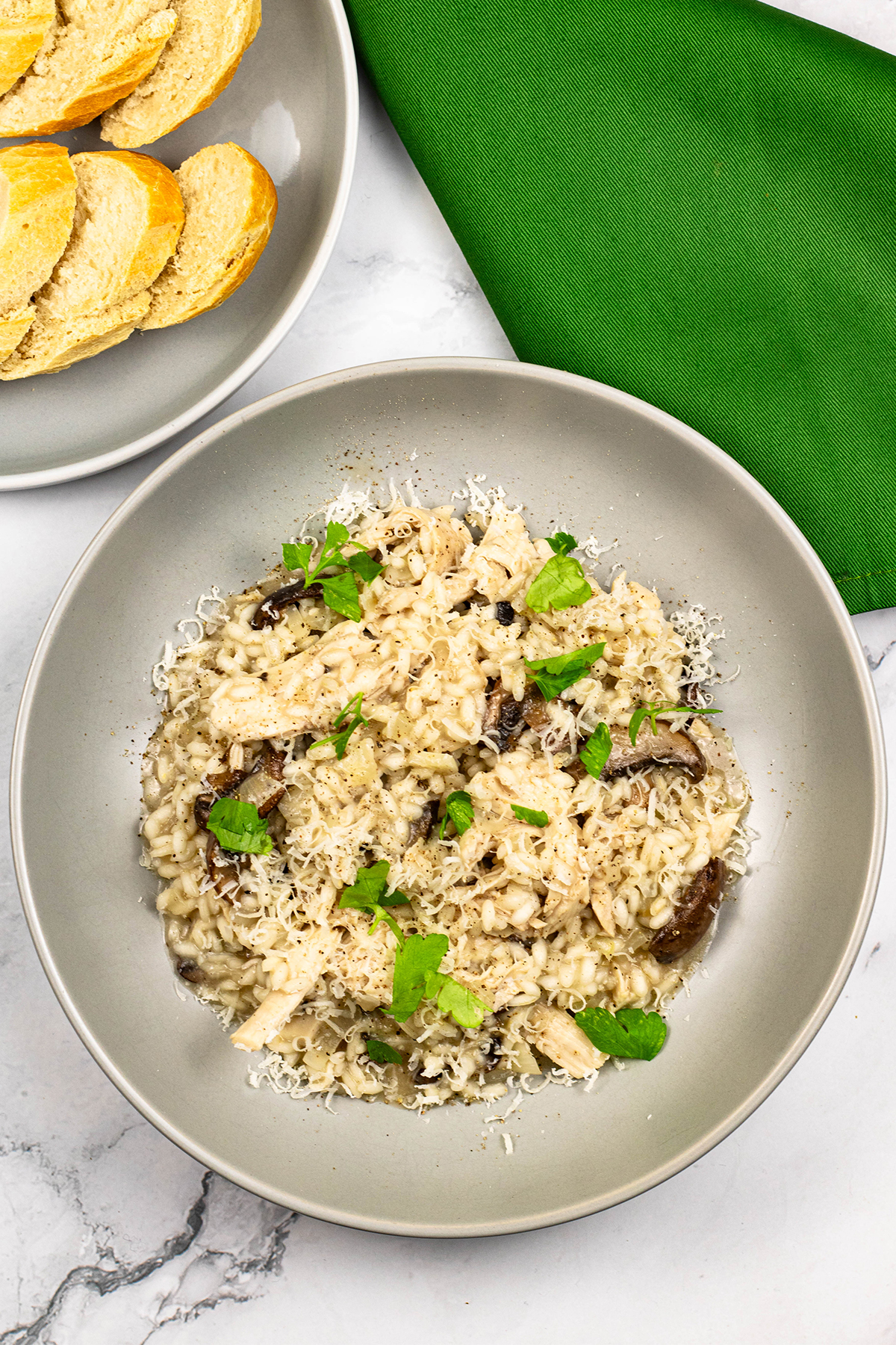 Chicken and mushroom risotto in a grey bowl with a plate of sliced crusty baguette and a green napkin on the side.