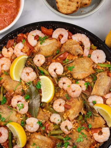 Chicken paella with prawns and beans in a paella pan with dishes of pan con tomate and toasted crusty bread on the side.