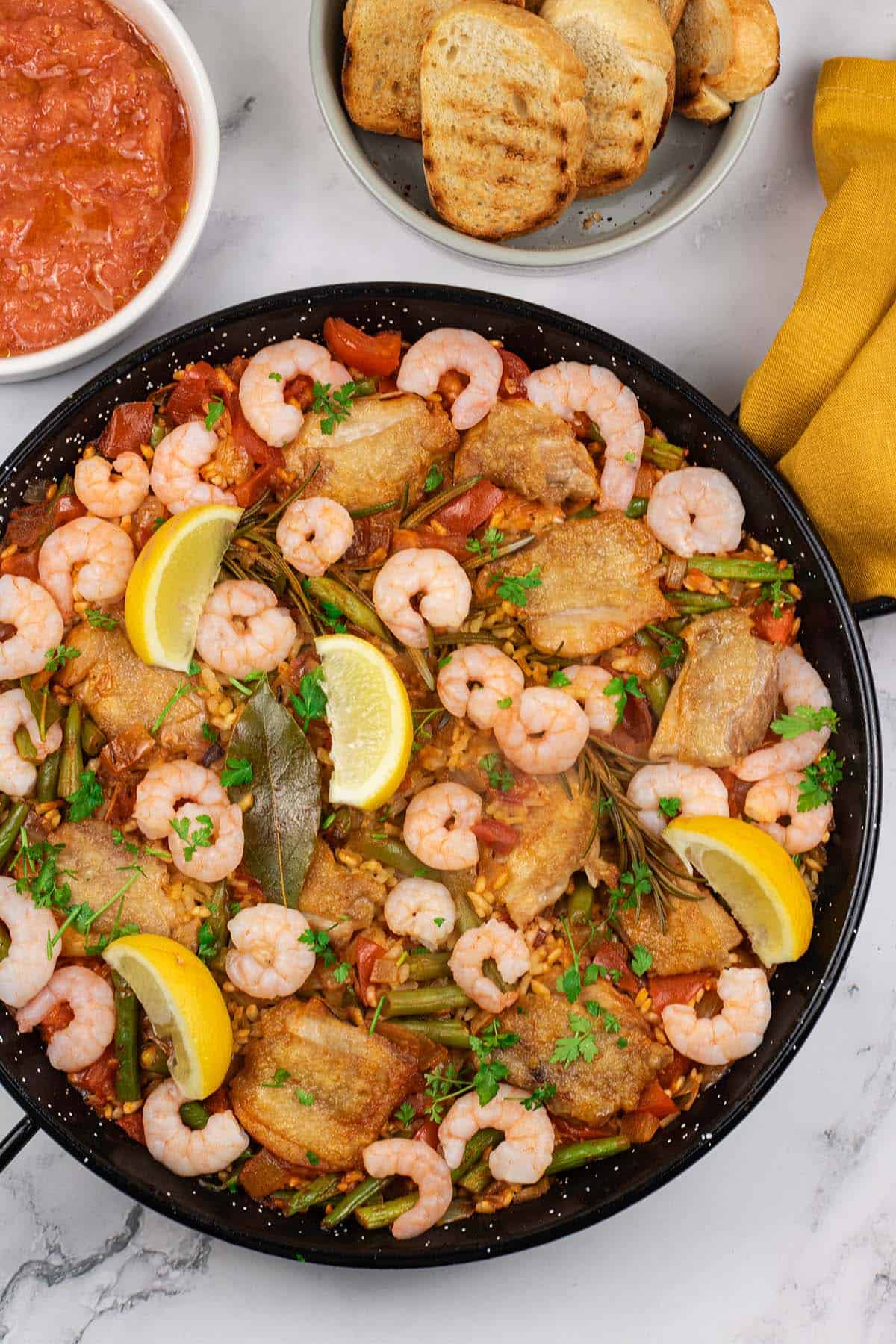 Chicken paella with prawns and beans in a paella pan with dishes of pan con tomate and toasted crusty bread on the side.