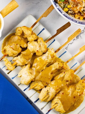 Chicken skewers with satay sauce on a striped platter, with a jug of sauce and bowl of vegetable fried rice on the side.