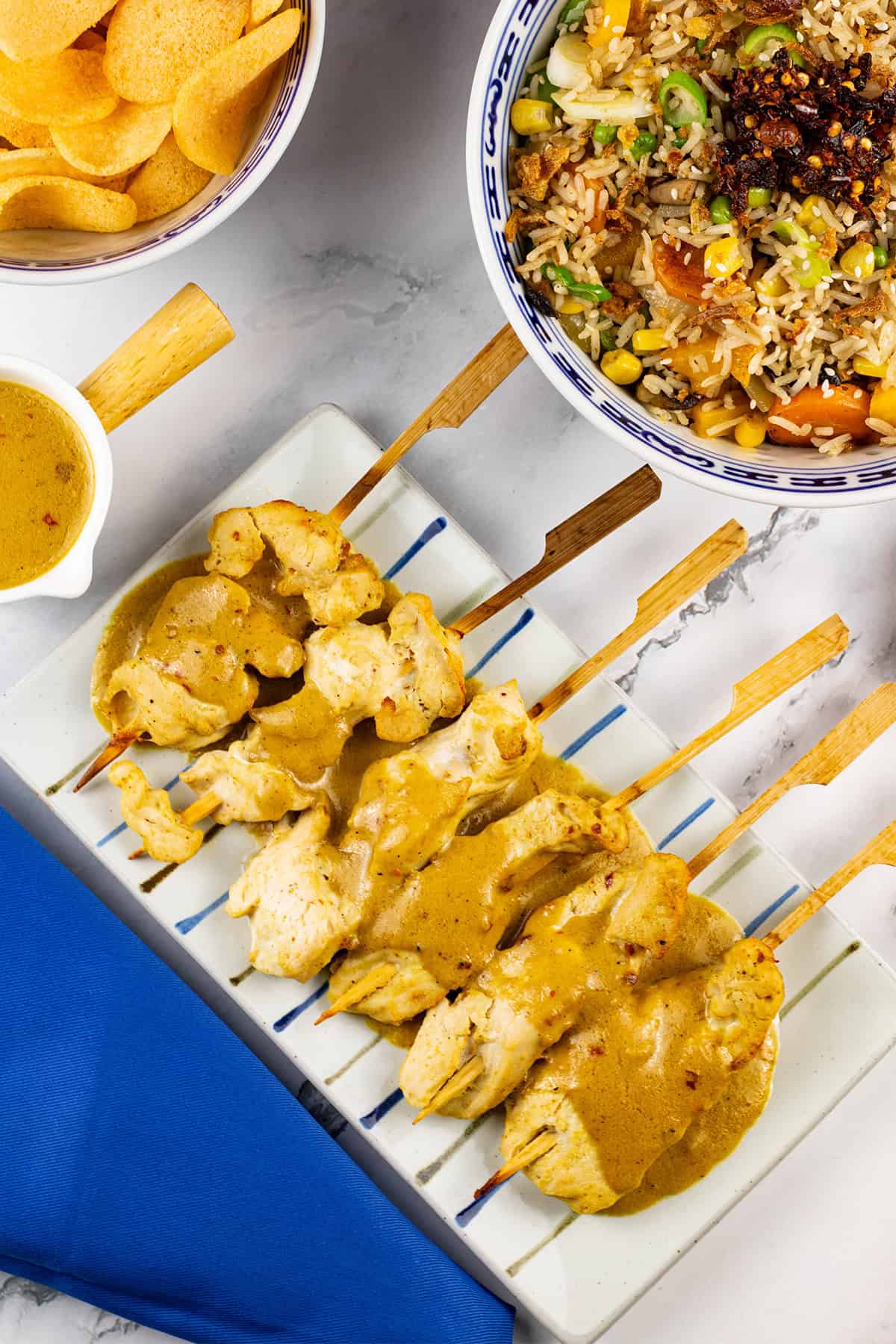 Chicken skewers with satay sauce on a striped platter, with a jug of sauce and bowls of vegetable fried rice and prawn crackers on the side.