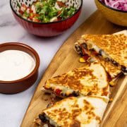 2 portions of chipotle black bean and corn quesadillas on a board with bowls of pickled red onions, pico de gallo salsa and sour cream on the side.
