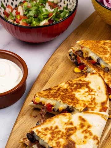 2 portions of chipotle black bean and corn quesadillas on a board with bowls of pickled red onions, pico de gallo salsa and sour cream on the side.