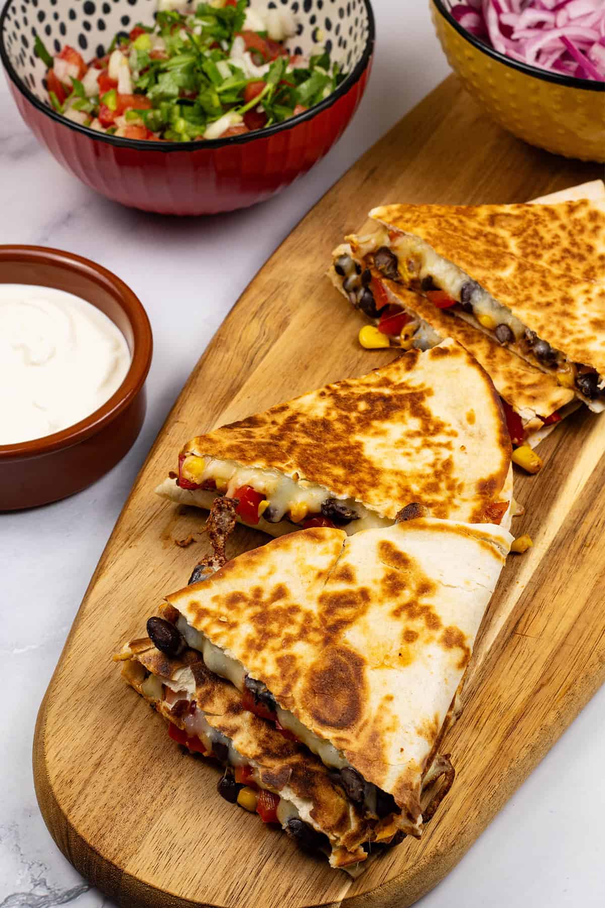 2 portions of chipotle black bean and corn quesadillas on a board with bowls of pickled red onions, pico de gallo salsa and sour cream on the side.