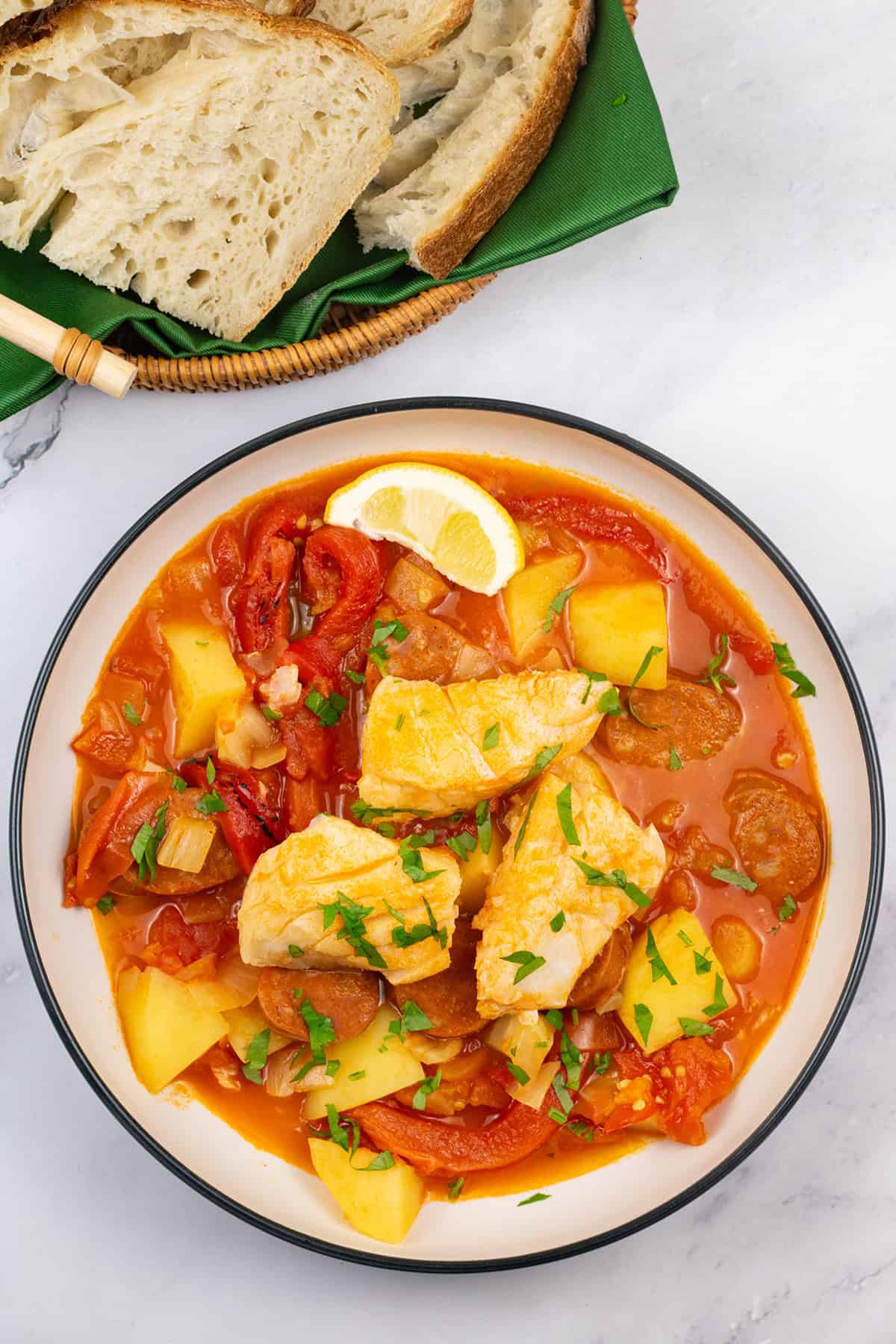 A portion of cod and chorizo stew in a bowl with a basket of crusty bread on the side.