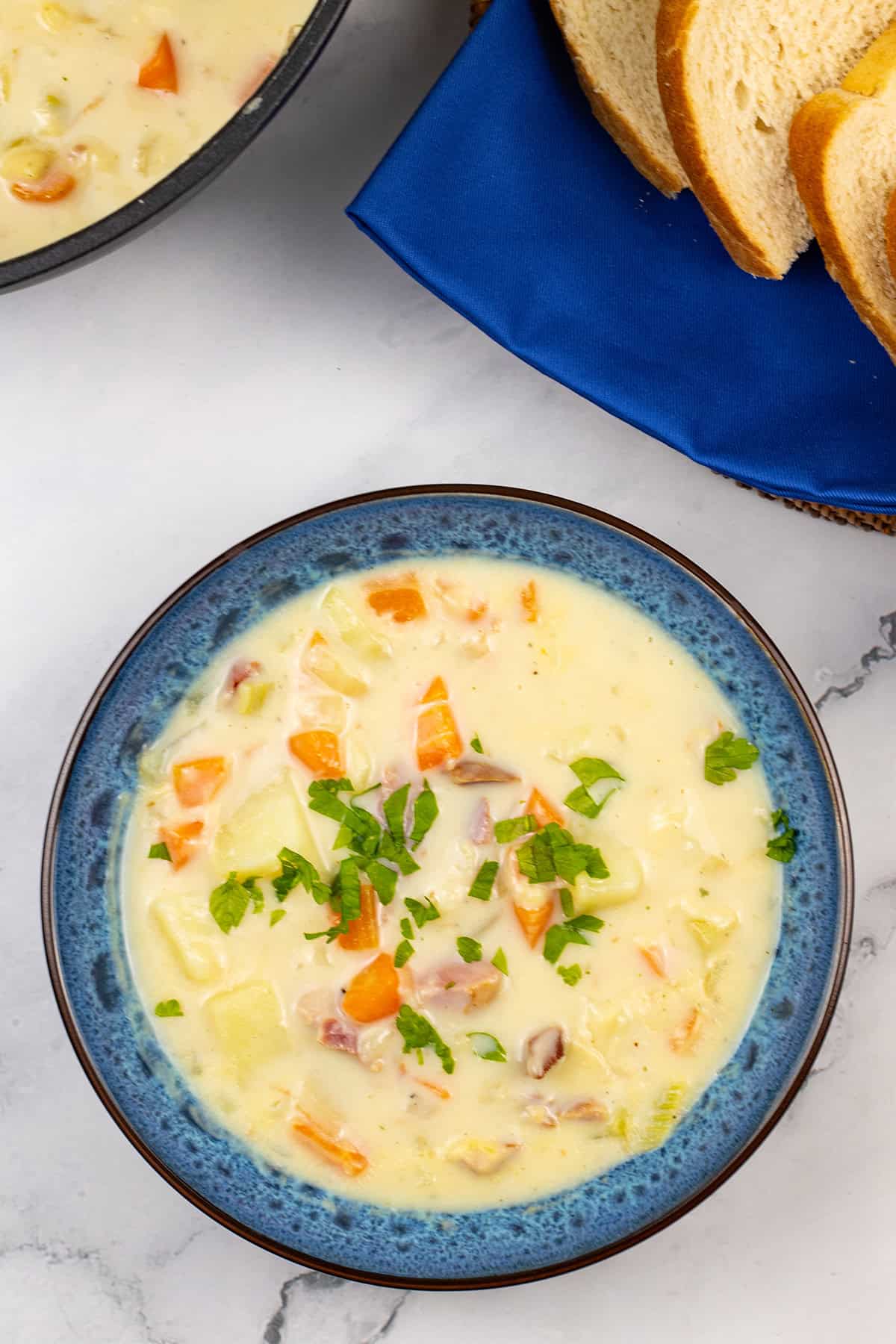 Creamy ham and potato soup in a blue patterened bowl with crusty bread and pot of soup on the side.