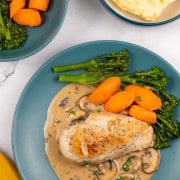 A portion of creamy mushroom chicken with broccoli and carrots on a teal dinner plate with bowls of broccoli and carrots, and mashed potatoes on the side.