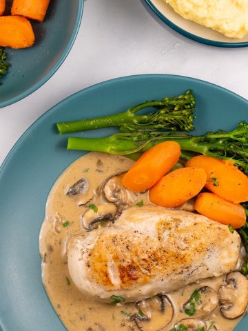 A portion of creamy mushroom chicken with broccoli and carrots on a teal dinner plate with bowls of broccoli and carrots, and mashed potatoes on the side.