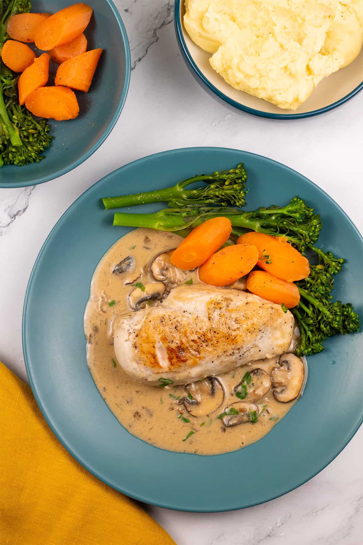 A portion of creamy mushroom chicken with broccoli and carrots on a teal dinner plate with bowls of broccoli and carrots, and mashed potatoes on the side.