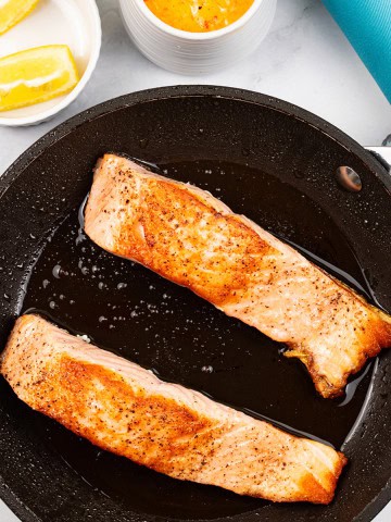 Crispy pan-fried salmon fillets in the frying pan with lemon wedges and a small jug of red pepper and paprika hollandaise on the side.