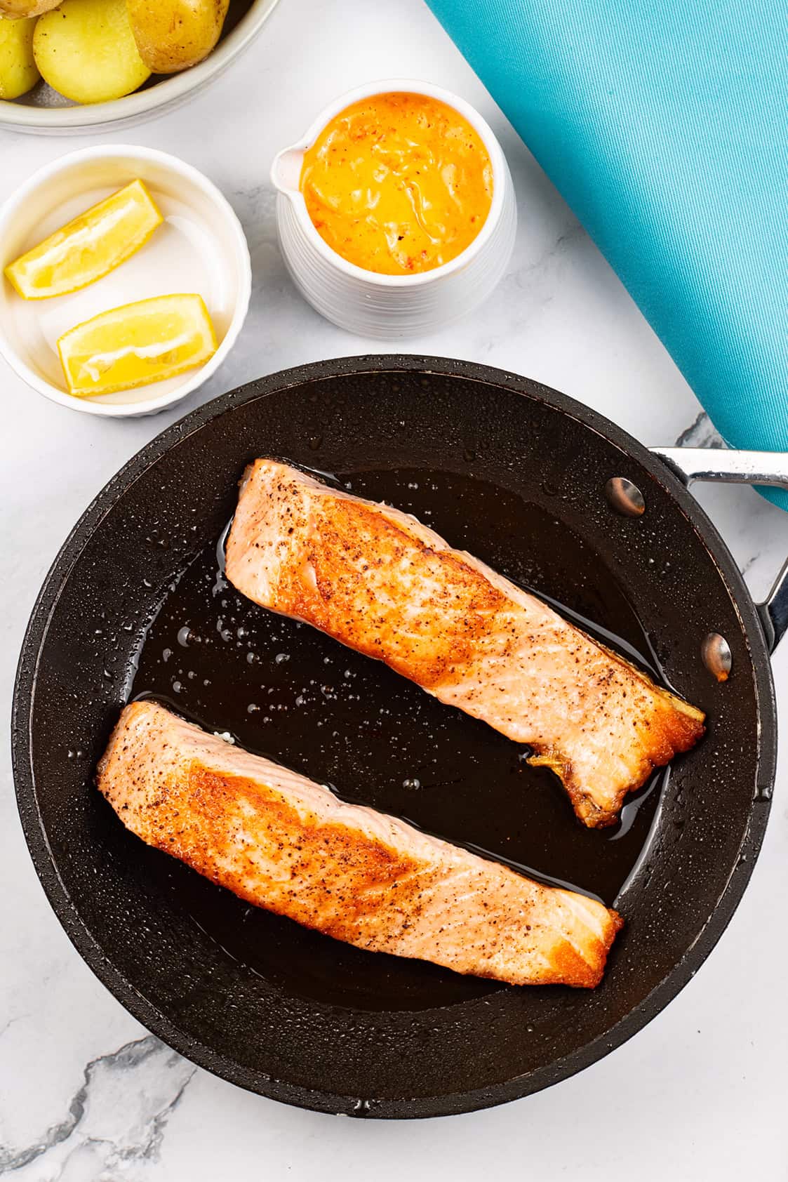 Crispy pan-fried salmon fillets in the frying pan with lemon wedges, baby potatoes and a small jug of red pepper and paprika hollandaise on the side.