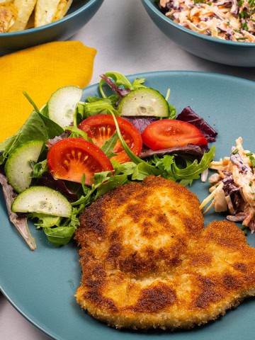 A crispy pork schnitzel, coleslaw and salad on a teal dinner plate, with a mustard napkin and bowls of chips and coleslaw on the side.