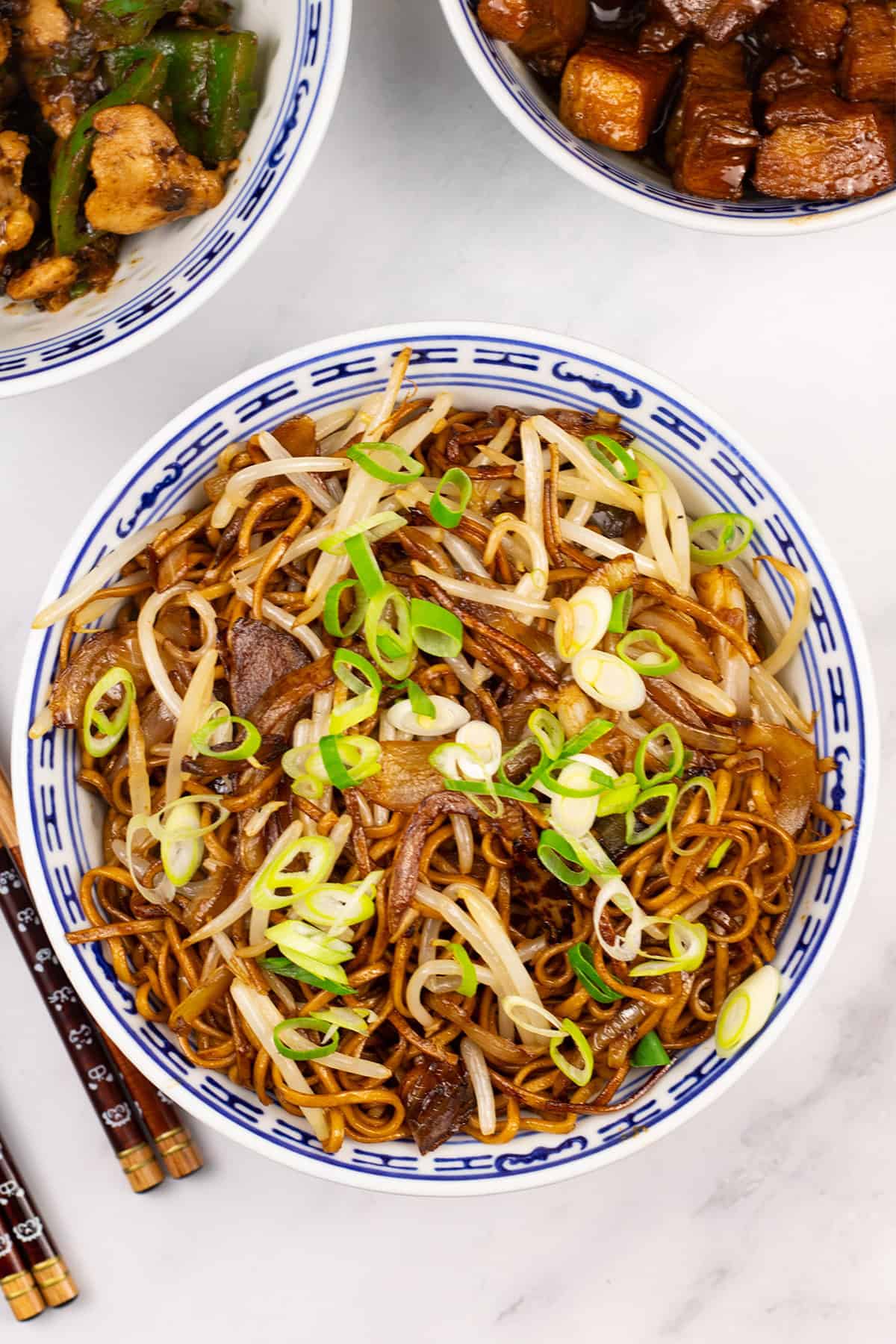 Fried noodles with beansprouts in an oriental bowl with bowls of chicken and green peppers and Shanghai pork, and chopsticks on the side.