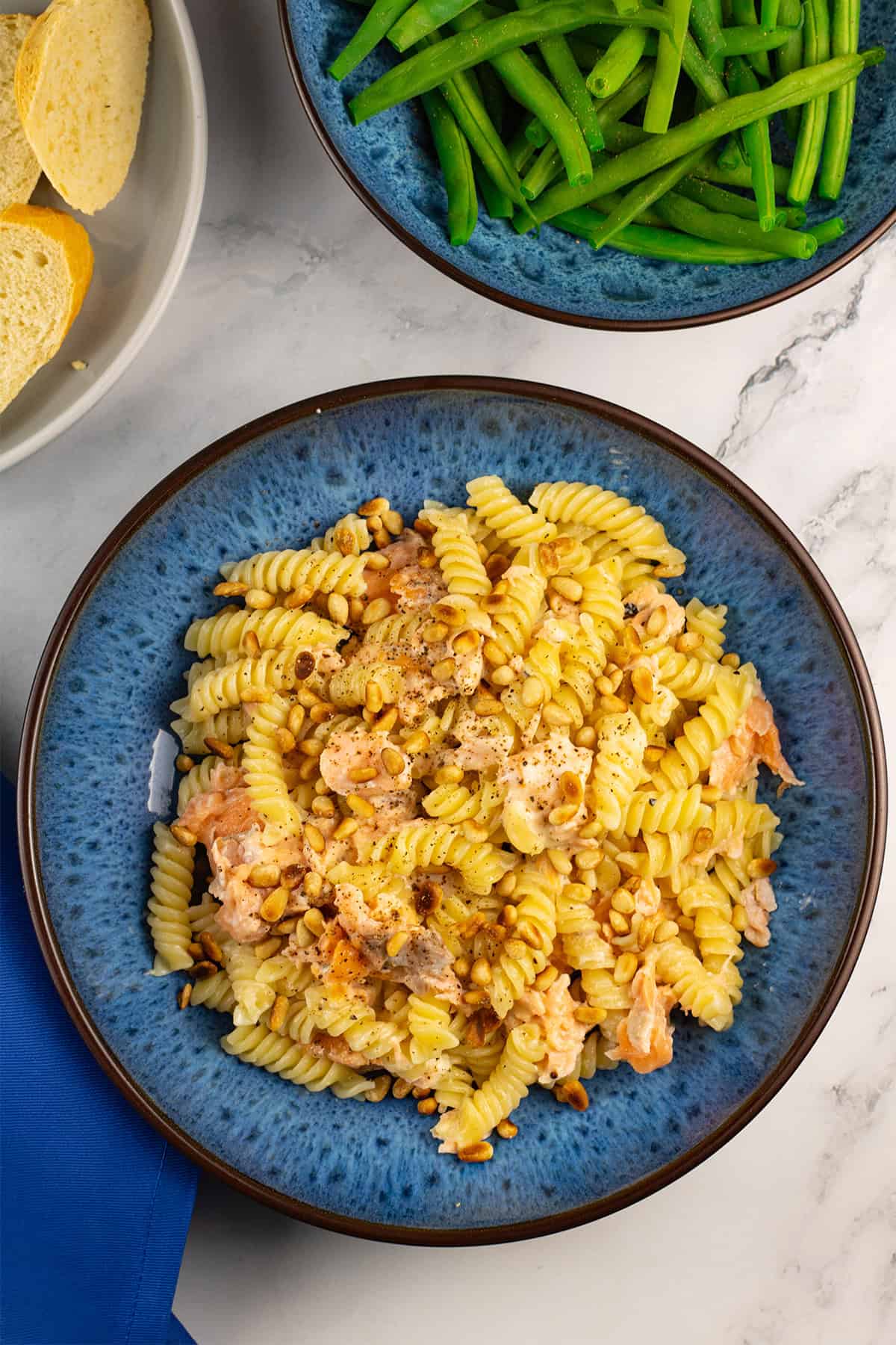 Hot smoked salmon with creamy pasta and pine nuts in a blue patterned bowl, with a bowl of green beans and plate of crusty bread on the side.